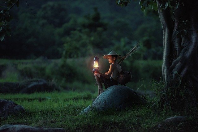 Dieťa sedí na kameni pri strome a v ruke drží zapálený lampáš.jpg