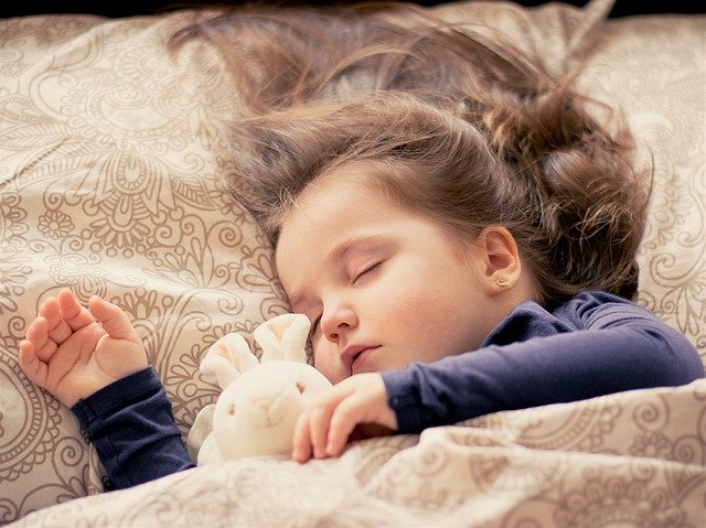 Dievčatko s dlhými vlasmi spí s medvedíkom v posteli pri svetle.jpg